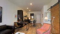This single-floor space showcases an open-concept layout that efficiently combines living, dining, and kitchen areas into one cohesive environment. The kitchen is outfitted with dark espresso cabinetry, stainless steel appliances including a full-size refrigerator and microwave, and a functional countertop workspace. A compact kitchen island with bar-height seating on casters serves as both a dining and casual gathering area, offering flexibility in how the space is used.

The living area features warm-toned upholstered seating, adding a welcoming contrast to the darker kitchen cabinetry. A wooden desk with an industrial-style metal frame and a leather chair define a dedicated workspace area, making this space well-suited for those who work from home. Light hardwood flooring runs throughout, contributing to a bright and cohesive aesthetic. Recessed ceiling lighting provides consistent illumination across the open floor plan. The white walls and neutral tones keep the space feeling clean and airy, while thoughtful furniture placement maximizes functionality without overcrowding the layout.