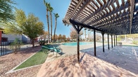 Located at 4870 Nellis Oasis Ln in Las Vegas, NV, this outdoor recreational space offers a well-appointed community amenity area designed for leisure and relaxation under the Nevada sun. The centerpiece of the space is a generously sized swimming pool with a smooth curved design, finished in clear turquoise water and surrounded by a wide concrete pool deck ideal for sunbathing and socializing. Stainless steel entry rails provide safe access into the pool. Surrounding the pool, patches of maintained artificial turf are accented with desert-friendly landscaping, including decorative red gravel, shrubs, and an array of mature palm trees and deciduous trees that provide natural shade and visual interest. Teal lounge chairs and seating are positioned throughout the green areas, offering comfortable spots to unwind. A chain-link-enclosed tennis court is situated adjacent to the pool area, complementing the recreational offerings. Tall light poles are installed throughout, ensuring the space is functional during evening hours. The community buildings visible in the background feature a Southwestern-inspired architectural style with stucco exteriors, red tile roofing, and bold accent colors. The overall ambiance is bright, open, and inviting, characteristic of a well-maintained Las Vegas residential community.