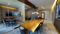 This single-floor professional office space combines modern design with refined materials to create a polished and functional work environment. The centerpiece of the space is a stunning live-edge wood conference table, finished in a warm natural tone and surrounded by sleek contemporary chairs, making it ideal for meetings and collaborative sessions. Suspended above the table, a pair of elongated linear pendant lights cast a warm, focused glow that highlights the table's natural grain and anchors the room visually.

Glass-partitioned offices line the perimeter, offering transparency and openness while maintaining acoustic separation for focused work. Dark wood framing accents the glass panels, adding warmth and structure to the otherwise modern aesthetic. The private offices within are furnished with ergonomic seating and curated artwork, lending a professional yet inviting atmosphere.

The surrounding walls feature exposed concrete textures, contributing an industrial character that is balanced by recessed ceiling lighting and a neutral-toned carpet with a subtle geometric pattern. A dedicated service or print station is visible near a solid dark-paneled door, ensuring practical functionality is seamlessly integrated into the design. Overall, the space strikes a compelling balance between executive sophistication and contemporary workplace design.