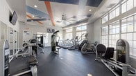 This fitness center is a thoughtfully designed, single-floor facility that offers a comprehensive workout experience. The space is defined by its striking tray ceilings accented with bold blue and orange geometric panels, complemented by large, multi-pane windows that line the walls and flood the interior with abundant natural light. An oversized ceiling fan anchors the main workout area, enhancing airflow throughout the space.

The cardio zone is well-appointed with a row of treadmills, elliptical machines, and a recumbent bike, all positioned near the windows to maximize natural light and outdoor views. Adjacent to this area, a dedicated strength training section houses cable machines, a dumbbell rack, and a variety of weight benches. A separate functional fitness zone features cycling bikes, medicine balls, resistance bands, battle ropes, and exercise mats, supporting group and circuit-style training.

The facility also includes a convenience station with white cabinetry, a granite countertop, a stainless steel sink, and a built-in beverage cooler — ideal for post-workout refreshment. The durable rubber flooring throughout provides both safety and comfort. Wall-mounted televisions and mirrored surfaces further enhance the workout environment, making this fitness center a fully equipped and welcoming space for all fitness levels.