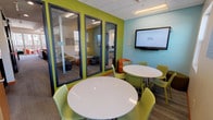 This energetic collaborative space is thoughtfully designed to support both group study and individual focus. The main area features two round white tables surrounded by lime green perforated chairs, creating an inviting and functional environment for group work or discussion. A large wall-mounted flat-screen display is positioned on a soft blue accent wall, making the space well-suited for presentations and collaborative projects. The interior design draws on a bold, modern color palette, with vibrant lime green walls contrasting against the calming blue tones, lending the space a lively yet focused atmosphere. Carpet tile flooring and a drop ceiling with recessed lighting complete the practical, well-lit environment. Along one wall, floor-to-ceiling glass-paneled private study rooms offer quieter, enclosed spaces for individuals or small groups, maintaining visual openness while providing acoustic separation. Natural light pours in through multiple windows that frame scenic mountain views and the surrounding landscape, adding a refreshing outdoor connection to the interior. A unique natural wood stump accent piece adds a touch of organic warmth to the otherwise contemporary setting. Study room policies are clearly posted, indicating this space is designed for structured, shared use within a larger institutional or educational facility.
