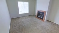 This single-story space includes a living area finished with neutral wall-to-wall carpeting and light gray painted walls, creating a clean and understated backdrop. A standout feature of the room is the fireplace, framed by a terra cotta-toned tile surround and a dark metal facade, adding a focal point and functional warmth to the space. A large window fitted with horizontal blinds faces the street, allowing natural light to fill the room while offering views of the surrounding exterior. The overall ambiance is simple and move-in ready, with a neutral palette that provides a versatile foundation for a variety of interior styles.
