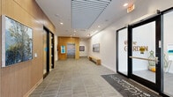 The Residences at the Garden offers a thoughtfully designed single-floor amenity space that blends contemporary architecture with warm, welcoming interiors. The main entrance lobby sets an immediate tone of sophistication, featuring large-format tile flooring, warm wood-paneled walls, recessed linear ceiling details, and stainless steel elevator doors framed by curated artwork. A branded entry mat and floor-to-ceiling glass doors complete the polished arrival experience.

Beyond the lobby, the amenity lounge opens generously, anchored by a statement linear pendant light fixture and expansive floor-to-ceiling glazing that frames views of a lush outdoor garden and patio. The space transitions fluidly between a comfortable seating area with upholstered lounge chairs and a well-appointed kitchenette outfitted with light wood cabinetry, open shelving, a microwave, and a coffee station. A media area with tile flooring features a large flat-screen television atop a dark display cabinet, accented by a gallery wall of framed black-and-white photography.

A private meeting room provides a focused workspace, highlighted by a sculptural hanging art installation, pendant lighting, and a collaborative table with ergonomic seating. Throughout, an art-forward aesthetic prevails, with textile works, photography, and mixed-media pieces displayed across the space, contributing to a refined yet inviting community atmosphere.