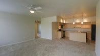 This single-floor residence features an open-concept layout that seamlessly connects the living area to the kitchen, creating a spacious and functional flow. The living room is finished with neutral wall-to-wall carpeting and includes a ceiling fan with an integrated light fixture, providing both comfort and practical illumination.

The kitchen is positioned along the back wall and is equipped with light maple cabinetry offering ample storage space. Black appliances, including a range and microwave, contrast cleanly against the cabinetry. A peninsula with a raised breakfast bar separates the kitchen from the living area, adding a casual dining option and defining the two spaces without closing them off. A pendant light hangs above the peninsula, complementing the track lighting installed along the kitchen ceiling for targeted task lighting. A sink is positioned along the counter, completing the functional kitchen layout.

A hallway visible from the living area leads to additional rooms, suggesting a well-organized floor plan. The neutral color palette, consistent flooring, and clean lines throughout contribute to a bright, cohesive, and move-in-ready atmosphere.