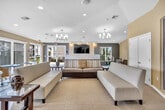 Located in Murrieta, CA, this single-floor community clubhouse offers a thoughtfully designed layout that balances comfort and functionality. The open living room serves as the heart of the space, featuring elegant dual chandeliers, recessed lighting, and large windows that flood the room with natural light. Plush seating arrangements are anchored by a patterned area rug over light tile flooring, while a wall-mounted flat-screen TV above a sleek fireplace with a stone surround creates a welcoming focal point. Glass doors open directly to an outdoor pool area, seamlessly blending indoor and outdoor living. The kitchen is a standout feature, showcasing rich dark wood cabinetry, a striking multicolored mosaic tile backsplash, stainless steel appliances including a double oven and full-size refrigerator, and a curved peninsula with a neutral countertop and modern bar seating. A stainless steel island range hood adds an industrial-chic touch. The separate dining room offers an intimate setting with warm neutral tones, carpeted flooring, and understated wall art. Throughout the space, warm golden-yellow accent walls contrast with crisp white ceilings, contributing to a bright, inviting ambiance ideal for gatherings and community events.