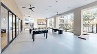 Located at Meritum Sonoran Desert in Phoenix, AZ, this single-floor amenity lounge offers a generously sized, open-concept space designed for community recreation and social gathering. The interior is defined by a clean, contemporary aesthetic featuring smooth concrete flooring, neutral stucco-finish walls, and high ceilings fitted with sleek black ceiling fans that keep the space comfortable year-round.

The layout is highly versatile, incorporating a dedicated dining area alongside a recreation zone equipped with a foosball table, ping pong table, and a large-format connect-four game. Comfortable lounge seating with soft mint-green cushions provides relaxed gathering spots throughout. A wall-mounted flat-screen TV and a striking moss-wall art installation bearing the Meritum Sonoran Desert branding serve as standout focal points, adding both visual interest and a touch of nature-inspired design.

Large open-air cutouts and floor-to-ceiling sliding glass doors flood the space with abundant natural light while offering direct visual connection to the surrounding desert-inspired landscaping, complete with native plants and cacti. The seamless transition between the indoor and semi-outdoor covered lounge areas creates an inviting, airy atmosphere that reflects the spirit of Sonoran Desert living. The entrance is anchored by a welcoming branded entry mat and a curated pair of accent chairs.