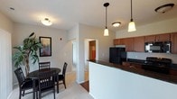This single-floor residence features an open-concept layout that connects the kitchen and dining area, creating a cohesive and functional living environment. The kitchen is equipped with rich medium-toned wood cabinetry, dark granite-style countertops, and black appliances, including a full-size refrigerator and microwave. A peninsula with a raised breakfast bar extends into the living space, serving as both a prep area and a natural divider between the kitchen and dining zone. Pendant lights with warm-toned shades hang above the peninsula, complementing the flush-mount ceiling fixtures throughout.

The dining area accommodates a round dark-wood table with matching chairs, positioned on plush carpeting that adds warmth underfoot. The transition from carpet to hardwood-style flooring marks the shift between spaces, adding visual interest. Neutral-toned walls provide a clean, versatile backdrop throughout. A hallway visible from the main living area leads to additional rooms, suggesting a well-organized floor plan. The overall ambiance is warm and functional, with thoughtful design details that balance practicality and comfort.