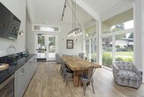 Located at 103 Felix St in Santa Cruz, California, this single-floor space offers a well-appointed, versatile gathering environment with a clean, modern-rustic aesthetic. The interior is defined by vaulted, beam-accented ceilings with white shiplap detailing that lend a sense of openness and volume throughout.

The main gathering area features expansive floor-to-ceiling windows along one wall, flooding the space with abundant natural light and offering unobstructed views of a landscaped outdoor area, pool, and mature trees beyond. Light hardwood flooring runs throughout, complementing the neutral warm-gray wall palette. Seating includes patterned upholstered accent chairs paired with sleek black side tables, alongside a rustic wood communal dining table with industrial-style pendant lighting overhead.

A built-in bar area runs along one interior wall, outfitted with white shaker cabinetry, a polished dark granite countertop, a stainless steel beverage cooler, and a wall-mounted flat-screen television flanked by vintage-style sconce lighting. An accessible restroom is conveniently located adjacent to this service area.

Double glass entry doors open directly onto a wood deck, creating a seamless indoor-outdoor connection. The overall ambiance is inviting, polished, and well-suited for community gatherings or private events.