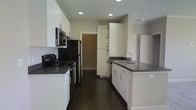 This single-floor space centers around a well-appointed kitchen that combines functionality with a clean, modern aesthetic. The kitchen is designed in a galley-style layout, featuring crisp white cabinetry that extends from floor to ceiling, providing ample storage throughout. Dark charcoal countertops create a striking contrast against the white cabinets, lending the space a contemporary feel. Stainless steel appliances, including a gas range, over-the-range microwave, and refrigerator, line one wall, while a double-basin sink with a sleek faucet is positioned along the opposite counter. A tall pantry-style cabinet adds additional storage near the passageway. Recessed lighting in the ceiling provides consistent, even illumination throughout the kitchen. The dark hardwood flooring anchors the space and complements the overall color palette, running the length of the room. A doorway visible at the end of the kitchen connects to an adjacent area, suggesting a practical, open flow between spaces. The overall ambiance is clean, minimal, and functional.