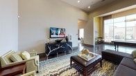 This single-floor communal space is designed as a contemporary social hub, blending recreational and lounge amenities within an open, airy layout. Soaring ceilings throughout create a grand sense of volume, while polished concrete floors lend an industrial-modern aesthetic that ties the entire space together.

The game area is anchored by a full-size billiards table positioned beneath a statement pendant light fixture, bathed in abundant natural light streaming through dramatic floor-to-ceiling windows. These expansive glazed panels frame views of the surrounding neighborhood and draw sunlight deep into the interior. Sheer drapery panels frame the windows, adding softness to the otherwise sleek architecture.

Adjacent to the game area, a well-appointed lounge zone features a large sectional sofa, coordinating accent chairs, and a bold chevron-patterned area rug. A wall-mounted flat-screen television and a sculptural media console with decorative branch detailing serve as focal points. Colorful artwork on accent walls painted in warm yellow tones adds visual interest throughout.

A kitchenette bar area offers a long granite-topped counter with bar seating, rich dark cabinetry, globe pendant lighting, and a stainless steel refrigerator — ideal for casual entertaining. The overall ambiance is modern, welcoming, and well-suited for social gatherings.
