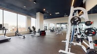 This contemporary fitness center is thoughtfully designed to support a full range of workout regimens in a polished, professional environment. The space is defined by dramatic dark ceilings accented with linear LED lighting and recessed fixtures, creating a sleek, high-energy atmosphere. Durable rubber flooring spans the entire facility, providing both safety and performance underfoot.

Floor-to-ceiling windows line multiple walls, flooding the space with natural light and offering expansive views of the surrounding cityscape — an energizing backdrop for any workout. The main training floor houses a comprehensive selection of free weights, including a full dumbbell rack and weight plates, alongside benches and cable machines to accommodate strength training of all kinds.

A dedicated cardio zone features a row of Precor ellipticals positioned directly against the glass perimeter walls, maximizing the outdoor views. Additional cardio equipment includes treadmills, stationary bikes, and a rowing machine. A boxing station with heavy bags and boxing gloves adds a functional training dimension to the space.

A glass-enclosed studio room with mirrored walls and TRX suspension trainers provides a private area for group classes or focused functional workouts. Full-length mirrors throughout the main floor further enhance the sense of openness and allow users to monitor their form. The overall design strikes a balance between functionality and modern aesthetics.