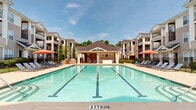 Located at 4625 Millbrook Green Dr in Raleigh, NC, this outdoor amenity space centers around a generously sized resort-style swimming pool with clearly marked lane lines and a shallow end designated for safety. The crystal-clear turquoise water is framed by a wide, sundrenched concrete pool deck furnished with rows of blue lounge chairs and colorful shade umbrellas, providing ample space for relaxation and recreation.

A charming covered cabana pavilion anchors one end of the pool area, featuring a hip-style shingled roof supported by decorative columns. The structure houses a bar counter and service area, making it well-suited for outdoor gatherings. Additional amenities, including what appears to be an outdoor grill station, are accessible along the perimeter of the deck.

The entire pool area is enclosed by a black iron fence, providing security while maintaining open sightlines to the surrounding landscaped grounds and mature trees. The multi-story residential buildings that frame the space feature classic architectural details including pitched rooflines, neutral siding, and private balconies. A separate entrance with brick detailing, broad concrete steps, and black metal railings provides a welcoming and accessible approach. Under clear North Carolina skies, this outdoor space offers a bright, inviting atmosphere ideal for leisure and community enjoyment.