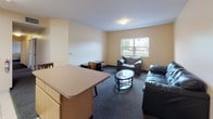 Located at 5086 Station Rd in Erie, Pennsylvania, this single-floor residential unit offers a functional and practical layout designed to accommodate multiple occupants. The shared living area features dark carpeting, leather seating, a glass-top coffee table, and a window that allows natural light and views of the surrounding buildings. Adjacent to the living space is a compact kitchen equipped with a stove, built-in microwave, stainless steel sink, and ample oak cabinetry providing generous storage above and below the countertops. Light tile flooring ties the kitchen area together cleanly. Multiple bedrooms are each furnished with a twin bed frame, a multi-drawer dresser, and carpeted flooring, offering a straightforward and comfortable personal space. One bedroom also features a mirrored sliding closet door, adding a sense of depth to the room. Individual bedroom doors are clearly marked, indicating a suite-style arrangement suited for shared living. Two full bathrooms serve the unit, each featuring light tile flooring, an oak vanity with under-sink storage, a wall mirror, and a shower-tub combination. The overall design is clean and utilitarian, emphasizing practicality and ease of use throughout.