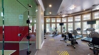 This single-floor fitness and recreation facility offers a comprehensive workout environment designed to accommodate a wide range of athletic activities. The main fitness area is elegantly appointed with tray ceilings, crown molding, and warm neutral-toned walls, creating an upscale atmosphere uncommon in gym settings. Decorative chandelier-style ceiling fans add a refined touch overhead. Large, multi-pane windows wrap around portions of the room, flooding the space with abundant natural light and providing views of the surrounding greenery — particularly notable in the curved treadmill alcove, where floor-to-ceiling windows create an immersive outdoor connection. The gym floor features patterned carpet tiles with bold yellow accent stripes, and the space is outfitted with a full suite of Precor and True cardio machines, including treadmills, ellipticals, and stationary bikes, as well as a multi-station strength training unit and free weights. Wall-mounted flat-screen televisions are positioned throughout for convenience. A full-wall mirror expands the sense of space in the strength area. Adjacent to the fitness room, a glass partition separates a spacious indoor sports court with high ceilings, painted concrete block walls, red safety padding along the base perimeter, and a regulation basketball hoop, suitable for both basketball and racquetball.