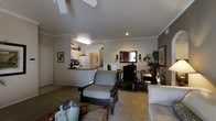 This single-floor residence offers a thoughtfully designed open-concept layout that seamlessly connects the main living areas. The living room is generously sized, featuring plush carpeting, crown molding, and a ceiling fan that adds both style and comfort. Tasteful framed artwork adorns the neutral-toned walls, contributing to a warm and inviting ambiance throughout.

The kitchen is efficiently laid out and visible from the main living space, featuring light wood cabinetry, a white refrigerator, and a raised breakfast bar that serves as a natural divider between the kitchen and the living area. The bar provides additional casual seating and counter space, making it ideal for both meal prep and entertaining.

Adjacent to the living room, a dedicated dining area accommodates a dark wood table and chairs set, accompanied by wall sconces that provide focused ambient lighting. A large mirror on the dining room wall enhances the sense of space and reflects natural light effectively.

Throughout the home, recessed lighting and wall sconces complement the natural light, creating a bright and comfortable environment. The consistent use of neutral tones, crown molding, and coordinated furnishings gives the space a cohesive, polished character well-suited for everyday living.