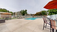 Located at 8015 36th Ave N in New Hope, Minnesota, this outdoor amenity area is centered around a generously sized, curved swimming pool with clear blue water and classic metal entry rails. The pool is set within a broad concrete sun deck that provides ample space for lounging, lined with cushioned chaise lounge chairs and accent umbrellas for shade. A white metal fence encloses the entire perimeter, offering both security and an open, airy feel that allows views of the surrounding mature trees and multi-story residential buildings beyond. On one side of the deck, a pergola structure anchors a dedicated outdoor seating and gathering area, complete with patio furniture and a charcoal grill, making it well-suited for social gatherings. Manicured landscaping, including a mix of deciduous and evergreen trees, adds natural beauty and a sense of privacy throughout the space. The overall ambiance is relaxed and community-oriented, designed to provide residents with a comfortable outdoor retreat for recreation and leisure during the warmer months.