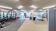 This expansive fitness center is thoughtfully designed to accommodate a wide range of workout disciplines across multiple interconnected zones. The space features a clean, modern aesthetic with white and neutral-toned walls, accented by bold pops of orange and slate blue, creating an energizing atmosphere throughout.

The main workout area is generously proportioned, with rubber flooring panels delineating a dedicated functional training and free-weight zone. This section is equipped with a multi-station cable machine, adjustable benches, a full dumbbell rack, medicine ball storage, and stability equipment. Floor-to-ceiling mirrors line the walls, enhancing the sense of openness and allowing users to monitor their form.

A separate cardio zone houses a row of treadmills and elliptical machines positioned directly in front of large windows, offering views of the surrounding cityscape and filling the area with natural light. Recumbent bikes and additional cardio equipment occupy a secondary area nearby.

The facility also includes a more intimate training room with rubber tile flooring, a power rack, free weights, and shelving for accessories — ideal for focused, small-group or personal training sessions. Bright linear fluorescent lighting illuminates every corner of the space uniformly. The overall layout is open and well-organized, designed to support both individual and group fitness routines efficiently.