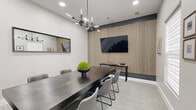 Located at 12167 Lincoln Lake Way in Fairfax, Virginia, this single-floor conference room is a thoughtfully designed professional meeting space that balances modern aesthetics with functional purpose. The room's most striking architectural feature is a floor-to-ceiling vertical wood slat accent wall set against a deep charcoal backdrop, which serves as the focal point and houses a large wall-mounted flat-screen display — ideal for presentations and collaborative sessions. A bold, multi-arm black chandelier anchors the ceiling, casting dynamic light across the space and complementing the room's contemporary design language. The centerpiece is a substantial dark-finished conference table surrounded by upholstered grey chairs with sleek metal frames, comfortably accommodating a full team. Patterned geometric carpeting in soft grey tones adds texture underfoot, while crisp white walls and roller shades on multiple windows allow for adjustable natural light. A narrow console table beneath the display provides additional surface space. Framed artwork and wall-mounted picture ledges contribute a curated, polished atmosphere. A large mirror on one wall enhances the sense of depth, and a glass-paneled black door connects the room to adjacent spaces, completing this refined and highly functional meeting environment.