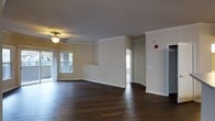 This single-floor residence offers a thoughtfully designed open layout that promotes a sense of spaciousness and ease of movement. The main living area is anchored by rich, dark hardwood flooring that extends throughout, providing a warm and grounded aesthetic. Elegant crown molding lines the ceilings, adding a refined architectural detail that elevates the overall interior finish. A ceiling fan with a light fixture is centrally positioned, contributing both functionality and understated style to the living space. Large windows along the exterior wall invite generous amounts of natural light, creating a bright and welcoming atmosphere. The walls are finished in a neutral, warm tone that complements the dark flooring and white trim work. A partial wall divider subtly defines separate areas within the open floor plan while maintaining visual continuity throughout the space. Doorways lead to additional rooms, suggesting a practical and connected layout. The combination of classic architectural details, quality flooring, and an open, light-filled design makes this residence a comfortable and visually cohesive living environment.