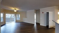 This single-floor residence offers a thoughtfully designed open layout that promotes a sense of spaciousness and ease of movement. The main living area is anchored by rich, dark hardwood flooring that extends throughout, providing a warm and grounded aesthetic. Elegant crown molding lines the ceilings, adding a refined architectural detail that elevates the overall interior finish. A ceiling fan with a light fixture is centrally positioned, contributing both functionality and understated style to the living space. Large windows along the exterior wall invite generous amounts of natural light, creating a bright and welcoming atmosphere. The walls are finished in a neutral, warm tone that complements the dark flooring and white trim work. A partial wall divider subtly defines separate areas within the open floor plan while maintaining visual continuity throughout the space. Doorways lead to additional rooms, suggesting a practical and connected layout. The combination of classic architectural details, quality flooring, and an open, light-filled design makes this residence a comfortable and visually cohesive living environment.