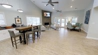 This single-floor space is designed as an open, inviting gathering area that combines functionality with a warm, coastal-inspired aesthetic. The soaring vaulted ceilings create an impressive sense of openness, enhanced by recessed lighting and dual ceiling fans that maintain airflow throughout. Light-toned wood-look flooring spans the entire space, contributing to its bright and airy feel.

A striking stacked-stone fireplace serves as the room's focal point, anchoring a comfortable lounge area furnished with neutral sofas, accent chairs in teal patterned fabric, and a coffee table arrangement ideal for casual conversation. A wall-mounted flat-screen television above the fireplace adds a modern entertainment element.

Toward one side, a tall bar-height table with gray upholstered stools provides an informal dining or work area. French doors along the far wall flood the space with natural light and offer views of the outdoor surroundings, while large windows further brighten the interior. The overall ambiance is relaxed yet polished, making this an ideal multi-purpose communal setting.