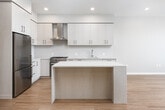 This contemporary single-floor residence showcases a clean, modern design with warm hardwood flooring that flows consistently throughout the open-concept living spaces. The kitchen is a standout feature, offering light wood flat-panel cabinetry extending to the ceiling, a marble-toned quartz countertop, a sleek stainless steel gas range with a metallic hood, a built-in wine cooler, and a generous island that doubles as a casual dining and prep surface. Recessed lighting illuminates the kitchen and living area, which opens toward expansive floor-to-ceiling windows that frame sweeping panoramic views, filling the space with abundant natural light.

The bedroom area is well-proportioned and connects to a custom-built walk-in closet featuring dark wood shelving, multiple hanging rails, and ample storage. A secondary bathroom offers a floating vanity with light wood cabinetry and a backlit mirror above a clean white sink, finished with neutral tile flooring. The primary bathroom is notably spacious, featuring a dual-sink floating vanity with a wide, illuminated mirror, a freestanding soaking tub, and a glass-enclosed walk-in shower, all finished in a refined, light neutral palette. The overall ambiance is polished and contemporary, with a cohesive design language throughout.