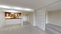 This single-floor residence is thoughtfully designed with an open, flowing layout that connects the main living area to the kitchen and private spaces beyond. The generous living room is finished with neutral wall-to-wall carpeting and is bathed in natural light from a wide bank of floor-to-ceiling sliding glass doors that span nearly the entire wall, opening onto a private patio. Mature trees visible through the glass create a serene, park-like backdrop that enhances the sense of privacy and connection to the outdoors.

The kitchen is positioned adjacent to the living area and features a pass-through counter with a rich, dark granite surface, warm honey-toned wood cabinetry with glass-front upper panels, and matching lower cabinets outfitted with stainless hardware. Appliances include a dishwasher and range, and the space is finished with light-toned tile flooring. A decorative frosted glass panel partition adds an architectural detail, subtly defining zones within the open plan without fully enclosing them.

A hallway leads to a carpeted bedroom that offers a quiet, private atmosphere with clean white walls and a closet accessible through a paneled door. Throughout the unit, neutral tones, consistent carpeting, and understated finishes create a cohesive, calm ambiance well-suited for comfortable everyday living.
