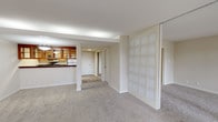 This single-floor residence is thoughtfully designed with an open, flowing layout that connects the main living area to the kitchen and private spaces beyond. The generous living room is finished with neutral wall-to-wall carpeting and is bathed in natural light from a wide bank of floor-to-ceiling sliding glass doors that span nearly the entire wall, opening onto a private patio. Mature trees visible through the glass create a serene, park-like backdrop that enhances the sense of privacy and connection to the outdoors.

The kitchen is positioned adjacent to the living area and features a pass-through counter with a rich, dark granite surface, warm honey-toned wood cabinetry with glass-front upper panels, and matching lower cabinets outfitted with stainless hardware. Appliances include a dishwasher and range, and the space is finished with light-toned tile flooring. A decorative frosted glass panel partition adds an architectural detail, subtly defining zones within the open plan without fully enclosing them.

A hallway leads to a carpeted bedroom that offers a quiet, private atmosphere with clean white walls and a closet accessible through a paneled door. Throughout the unit, neutral tones, consistent carpeting, and understated finishes create a cohesive, calm ambiance well-suited for comfortable everyday living.