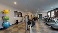 The Baxter fitness center is a comprehensively outfitted facility designed to support a full range of workout routines. The space features a distinctive checkered tile floor throughout, lending a clean and organized aesthetic that complements the modern, neutral-toned walls accented by deep teal and charcoal panels.

The main gym floor is generously sized, housing a robust selection of cardio equipment including multiple Matrix treadmills and ellipticals positioned along large windows that invite natural light and exterior views. A dedicated row of Hoist strength machines and cable systems lines one side of the room, while a well-stocked dumbbell rack with color-coded free weights serves the central floor area. Full-length mirrors expand the visual depth of the space and allow users to monitor their form during exercise. Ceiling fans and recessed lighting provide a comfortable environment throughout.

A separate room accessible through glass-paneled doors provides a quieter zone for focused training, featuring a stationary cycling machine, yoga mats, a wall-mounted television, and a water station. Wall-mounted racks neatly store stability balls in yellow, green, and blue, keeping the space tidy and functional. Together, these areas create a complete, amenity-rich fitness environment suited for both cardio-focused and strength-based training.