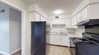 This single-floor unit offers a straightforward, functional layout with a clear separation between living and sleeping areas. The living room is a well-proportioned space featuring wood-style plank flooring and neutral gray walls, with a window fitted with horizontal blinds allowing natural light to filter in. An open passageway connects the living area directly to the kitchen, which is equipped with white cabinetry extending to the upper walls, a black refrigerator, a black electric range with coil burners, a stainless steel sink, and a range hood. Wood-style flooring continues throughout the kitchen, maintaining visual consistency with the main living area. The bedrooms feature soft, neutral-toned carpet and are each served by windows with blinds. One bedroom includes a standard closet, while another offers both a reach-in closet and an additional walk-in closet with built-in shelving, providing notable storage capacity. The bathroom features a full tub and shower combination enclosed with white ceramic tile. Throughout the unit, textured ceilings, white baseboards, and a consistent neutral color palette contribute to a clean, cohesive aesthetic.