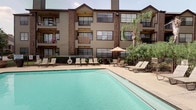 This outdoor pool area serves as a central amenity within a multi-story residential community, offering a well-appointed space for relaxation and recreation. The generously sized swimming pool features clear, blue-green water with metal handrail entry points and defined depth markings along the deck. Adjacent to the main pool is a attached spa, providing an additional option for leisure.

The surrounding concrete deck is furnished with multiple slatted lounge chairs and chaise lounges positioned for sunbathing, along with shaded seating areas beneath thatched-style umbrellas. A covered structure borders one side of the pool area, featuring dark wood post-and-beam detailing, stucco walls, and large glass sliding doors that open directly onto the deck. This covered walkway includes additional lounge seating and posted pool rules signage, providing a shaded transition space between the interior and pool deck.

Mature trees and tropical-style plantings, including palm trees, line the perimeter, adding greenery and privacy. The pool is enclosed by fencing with a safety ring station visible along one edge. The multi-story brick and siding building rises as a backdrop, with resident balconies overlooking the pool area.