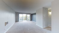This single-floor residence offers a well-organized layout with a cohesive neutral color palette accented by bold slate-gray feature walls throughout. The spacious living area is bathed in natural light, courtesy of large sliding glass doors that open onto a private balcony, offering pleasant outdoor views of surrounding greenery and neighboring buildings. Soft wall-to-wall carpeting extends throughout the main living spaces and bedrooms, contributing to a comfortable, understated ambiance. The kitchen is thoughtfully positioned adjacent to the living and dining area, featuring warm wood-tone cabinetry, dark countertops, and stainless steel appliances, including a dishwasher and gas range. A pendant light fixture adds a touch of character above the dining space. The bedroom is generously proportioned, highlighted by a dramatic dark accent wall and a built-in closet with sliding mirror doors, with a window that continues the theme of natural light. The unit includes two bathrooms: one with a walk-in shower enclosed by sliding glass doors and tile flooring, and a second featuring a full soaking tub with white tile surround and a grab bar, along with a small window for ventilation. Clean white cabinetry and classic fixtures complete the bathrooms' functional design.