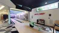 This single-floor entertainment space is designed around a private bowling alley that blends recreational functionality with bold, contemporary aesthetics. The lanes are finished in classic hardwood and equipped with a ball return mechanism, accompanied by a dedicated ball rack stocked with a colorful selection of bowling balls. A striking black-and-white photographic mural of the Great Wall of China spans the length of the lane wall, creating a dramatic visual backdrop that adds a sense of depth and grandeur to the space. On the opposite wall, a neon red script artwork adds a vibrant, artistic touch to the room's modern character. The ceiling is finished in a dark tone, contrasting cleanly with the white walls and contributing to the intimate, immersive atmosphere of the space. A large wall-mounted television is positioned above the lanes, ideal for scorekeeping or entertainment. A high, narrow clerestory window along the side wall introduces a strip of natural light, offering a glimpse of greenery outside. The combination of sleek finishes, artistic elements, and purpose-built amenities makes this a well-appointed recreational retreat.
