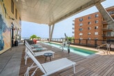 This rooftop amenity deck is a thoughtfully designed outdoor retreat that combines modern aesthetics with resort-style comfort. The centerpiece of the space is a sleek lap pool finished in cool blue tile, framed by warm wood decking that extends throughout the entire level. The pool is partially sheltered by a contemporary overhead canopy supported by striking angular concrete columns, providing shade while preserving open sightlines to the surrounding skyline and waterfront beyond the perimeter railing.

Adjacent to the pool, a dedicated sun lounge area features rows of white chaise lounges paired with small accent tables and an outdoor shower — perfect for relaxing after a swim. A bold, colorful mural of a woman dominates one exterior wall, adding an artistic and energetic focal point to this corner of the deck.

On the opposite end, a comfortable outdoor lounge area is anchored by a wall-mounted television and a sleek built-in fireplace, creating a cozy gathering space even on cooler evenings. Modern modular sofas in grey and orange complement the contemporary design language of the space.

Throughout the deck, panoramic views of the open water and surrounding urban landscape are visible, lending the space an airy, elevated ambiance. The combination of wood decking, concrete tile accents, and clean architectural lines gives this rooftop deck a polished, upscale character suited for both social gatherings and quiet relaxation.