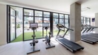 Located at 427 S Hill Street in the heart of downtown Los Angeles, this fitness center offers a well-appointed, single-floor facility designed for a comprehensive workout experience. The space features a clean, modern aesthetic with light hardwood-style flooring, bright recessed lighting, and expansive floor-to-ceiling windows that flood the interior with natural light and frame views of a lush green outdoor courtyard.

The cardio zone is equipped with multiple treadmills and stationary bikes, each outfitted with individual digital screens for on-demand entertainment and workout tracking. A dedicated functional training area houses a RAX multi-functional rig featuring TRX suspension trainers, kettlebells in various weights, medicine balls, a BOSU balance trainer, and resistance accessories — providing a versatile space for dynamic, full-body training.

The strength training floor offers an extensive selection of cable machines, weight racks, rowing machines, and resistance equipment arranged across a generously open layout. Full-length mirrors line the walls, enhancing the sense of space and aiding in form correction. Large flat-screen TVs are mounted throughout for added motivation.

A glass-partitioned area provides a separate exercise studio, while a dedicated service station with shelving offers towels and amenities. The overall ambiance is bright, airy, and professional, catering to a wide range of fitness goals.