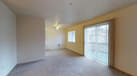 This single-floor space offers a clean, open layout designed for comfortable everyday living. The main living area is finished with wall-to-wall carpeting in a neutral tone, providing a soft and cohesive foundation throughout. Warm yellow-painted walls lend a welcoming brightness to the interior, complementing the natural light that pours in through multiple sources.

A large sliding glass door, dressed with full-length vertical blinds, serves as a prominent feature of the living area, opening to an outdoor space and offering views of the surrounding exterior. Adjacent to it, a standard window provides additional ventilation and light. A ceiling fan with a built-in light fixture is centrally positioned, offering both air circulation and overhead illumination.

The layout flows naturally between the main living zone and an adjoining area, separated by a partial wall that maintains an open feel while subtly defining separate functional spaces. A wall-mounted ventilation unit is also present, contributing to the room's climate control. The textured white ceiling adds a subtle visual detail overhead, while the overall design remains understated and versatile, providing a neutral canvas well-suited for a variety of furnishing styles and personal aesthetics.