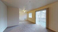 This single-floor space offers a clean, open layout designed for comfortable everyday living. The main living area is finished with wall-to-wall carpeting in a neutral tone, providing a soft and cohesive foundation throughout. Warm yellow-painted walls lend a welcoming brightness to the interior, complementing the natural light that pours in through multiple sources.

A large sliding glass door, dressed with full-length vertical blinds, serves as a prominent feature of the living area, opening to an outdoor space and offering views of the surrounding exterior. Adjacent to it, a standard window provides additional ventilation and light. A ceiling fan with a built-in light fixture is centrally positioned, offering both air circulation and overhead illumination.

The layout flows naturally between the main living zone and an adjoining area, separated by a partial wall that maintains an open feel while subtly defining separate functional spaces. A wall-mounted ventilation unit is also present, contributing to the room's climate control. The textured white ceiling adds a subtle visual detail overhead, while the overall design remains understated and versatile, providing a neutral canvas well-suited for a variety of furnishing styles and personal aesthetics.