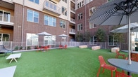 This outdoor dog park is a thoughtfully designed amenity space set within the courtyard of a contemporary multi-story brick building. The park is surfaced with vibrant, low-maintenance artificial turf that provides a clean and comfortable area for pets to roam freely. A secure perimeter of white metal fencing with latched entry gates ensures a safe, enclosed environment throughout the space.

Chunky natural stone bench blocks are positioned along one side of the park, offering rustic seating options for residents while their pets play. Scattered across the space are multiple round tables paired with bold red chairs and shaded by both striped and solid patio umbrellas, creating comfortable lounging spots for owners. Additional white bench seating with cushions lines the perimeter, further enhancing the social atmosphere of the area.

Slender young trees are planted along the fenced borders, introducing a touch of greenery and providing partial shade as they mature. The open-sky setting allows for abundant natural light throughout the day, and the surrounding building's brick facade and balconies frame the space with an urban residential character. The combination of practical pet-friendly features and well-considered human amenities makes this dog park a welcoming outdoor retreat.