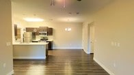 This single-floor residence offers a thoughtfully designed open-concept layout that effortlessly connects the kitchen, dining, and living areas into one cohesive, flowing space. Rich, dark hardwood flooring extends throughout, providing a warm and grounded aesthetic that complements the neutral, light-toned walls.

The kitchen is well-appointed with dark wood cabinetry, stainless steel appliances including a refrigerator, microwave, and gas range, and a central island that serves as both a prep area and a natural dividing point between the kitchen and the living space. Pendant lighting above the island adds a refined, modern touch.

A stylish chandelier anchors the dining area, casting a warm glow over the open space and adding an elegant focal point. The layout allows for generous room to arrange living and dining furniture with ease, making the space highly functional for both everyday living and entertaining.

A hallway visible from the main living area leads to additional rooms, each accessed through classic paneled doors. The overall ambiance is clean, contemporary, and welcoming, with a cohesive design language maintained throughout. Ample overhead lighting ensures the interior remains bright and inviting at all times.