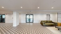 Located at 1435 51st St in North Bergen, NJ, this thoughtfully designed lobby and amenity floor offers a welcoming and stylish environment for residents. The single-floor layout encompasses a generously sized living area, a combined dining and living space, multiple hallways, and a dedicated package room, all connected by a cohesive modern aesthetic.

The main lounge area is a standout feature, showcasing a bold geometric three-dimensional patterned tile floor that anchors the room with visual interest. Two olive-green leather sofas flank an industrial-style coffee table with metal caster wheels, set atop a layered area rug. A large-scale decorative wall panel featuring delicate floral line art framed in a grid of black metal adds an artistic focal point to the room.

Black-framed glass double doors lead from the lounge into the dining area, which is bathed in natural light from a street-facing window offering views of the surrounding North Bergen neighborhood. A round white dining table with metal bistro-style chairs completes the space with a clean, contemporary touch.

The hallways are finished with smooth light-toned walls and recessed lighting, maintaining the building's sleek design language. A key-card-access package room with glass walls and polished stone flooring provides a secure and practical amenity. Throughout the space, crisp white ceilings with recessed lighting ensure a bright, well-lit atmosphere.