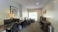 This single-floor space is designed as a functional and comfortable business center, thoughtfully arranged to accommodate a variety of work styles. The room features multiple built-in wooden desk workstations positioned along the walls, each equipped with ergonomic task chairs and personal computer setups, providing ample workspace for focused, individual tasks. Recessed ceiling lighting ensures consistent, even illumination throughout, while a centrally located window fitted with blinds allows natural light to filter in, creating a bright and welcoming atmosphere. The neutral, light gray wall tones paired with dark carpeted flooring give the space a professional yet approachable aesthetic. A comfortable seating area near the window, furnished with dark leather armchairs, offers a relaxed setting for reading or informal meetings. Decorative framed artwork adorns the walls, adding a touch of character and warmth to the otherwise utilitarian environment. The space is efficiently organized to maximize usability without feeling cramped, striking a balance between a professional office setting and an inviting, accessible environment suitable for a shared or community business center.