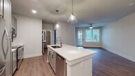 This single-floor residence presents a thoughtfully designed open-concept layout with a contemporary aesthetic carried consistently throughout. The kitchen serves as a central focal point, anchored by a large island with a white quartz countertop, an undermount sink with a pull-down faucet, and a built-in dishwasher. Sleek gray shaker-style cabinetry with matte black hardware lines the walls, complemented by a subway tile backsplash and a full suite of stainless steel appliances, including a refrigerator, range, over-the-range microwave, and pendant lighting overhead. Wood-look plank flooring flows through the kitchen and living area, which opens to a private balcony through a glass-panel door, filling the space with natural light. A ceiling fan adds both style and function to the living area. The bedrooms feature soft neutral carpet and windows with outdoor views. The bathroom is finished with wood-look flooring, a tub-shower combination with white tile surround, white quartz vanity top, and matte black fixtures. Multiple closets provide ample storage throughout.