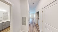 Located at The Cornerstone Westbury on Railroad Avenue in Westbury, NY, this thoughtfully designed single-floor unit offers a clean, modern aesthetic with high-quality finishes throughout. Light-toned hardwood-style flooring runs continuously across the space, complementing the crisp white walls and creating a cohesive, airy atmosphere.

The kitchen is a standout feature, outfitted with white shaker-style cabinetry extending to the ceiling for ample storage, bright white quartz countertops, and a full suite of stainless steel appliances including a refrigerator, range, built-in microwave, and dishwasher. A movable kitchen island with a matching quartz top adds flexible functionality to the space.

A hallway connects the main living area to the bedroom and bathroom, with a convenient electrical panel accessible along the corridor. The bedroom is a calm, neutral retreat featuring a window that allows natural light to filter in, with recessed ceiling vents ensuring year-round comfort.

The bathroom is elegantly appointed with marble-style floor-to-ceiling tile surrounding a soaking tub and shower combo, a white quartz vanity countertop, gray shaker-style cabinetry, brushed nickel hardware, and a large mirror — delivering a spa-like finish in a compact, efficient layout.
