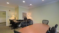 This single-floor office space offers a well-organized and professional environment designed to support productivity and collaboration. The room is finished with soft sage-green walls, crisp crown molding, and recessed lighting that provides even, comfortable illumination throughout the space. A central oval conference table surrounded by ergonomic mesh-back chairs serves as a focal point for meetings and collaborative work, while dedicated desk workstations along the perimeter are equipped with computers, task lighting, and ample surface area for individual tasks. The workstations are complemented by classic desk lamps that add a warm, inviting glow to the workspace. A window fitted with horizontal blinds allows for natural light control, contributing to a balanced and comfortable atmosphere. The neutral-toned carpeted flooring ties the room together, creating a cohesive and professional aesthetic. Overall, this office strikes a balance between a focused work environment and a collaborative meeting space, making it well-suited for small teams or shared office use.