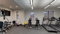 The fitness center at 101 Bill Robinson Way in Newburgh, NY is a well-appointed, single-floor facility designed to support a full range of workout routines. The space features durable rubber flooring throughout, providing both safety and comfort during exercise. Bright linear fluorescent lighting, supplemented by recessed ceiling lights, ensures the entire area is evenly and thoroughly illuminated.

The cardio section is equipped with multiple treadmills, recumbent bikes, upright stationary bikes, elliptical machines, and a rowing machine, offering users a wide variety of aerobic training options. A stability ball rounds out the functional fitness offerings. The strength-training area features a multi-station cable machine and a leg extension/curl attachment, providing options for resistance and weight training. A wall-mounted flat-screen television is positioned in the strength area for entertainment during workouts.

Multiple windows line the walls, allowing natural light to filter into the space and providing views of the surrounding area. The neutral gray walls and clean white trim create a modern, distraction-free environment conducive to focused exercise. A private room with glass panel doors is accessible from the main floor, offering additional utility. The overall layout is open and organized, making efficient use of the available space while ensuring comfortable movement between equipment stations.