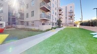 Located at 18750 Delaware St in Huntington Beach, California, this outdoor amenity space is a thoughtfully designed ground-level courtyard set within a contemporary multi-story residential community. The space is defined by expansive stretches of vibrant artificial turf that create a clean, well-maintained green landscape throughout. Wide concrete pathways cut through the lawn areas, providing easy circulation and connecting the various activity zones across the courtyard.

Seating is distributed generously throughout the space, including bold yellow benches and lightweight metal chairs that offer casual gathering spots along the perimeter. A dedicated lawn games area features cornhole boards, adding a recreational element to the outdoor experience. At one end of the courtyard, a BBQ and lounge zone is outfitted with bright red Adirondack chairs and an outdoor grill structure, making it well-suited for social gatherings.

Mature trees and manicured hedgerows line portions of the boundary walls, providing natural privacy screening and shade. The surrounding multi-story residential building features balconies overlooking the courtyard, reinforcing the sense of an active, community-oriented outdoor environment. Under the abundant Southern California sunshine, the space feels open, energetic, and inviting — a well-appointed outdoor retreat in the heart of Huntington Beach.