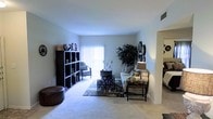 This single-story space features a generously sized living area finished with wall-to-wall carpet that lends a soft, comfortable feel underfoot. The room is anchored by a sliding glass door dressed with vertical blinds, allowing natural light to filter in and offering a connection to the outdoors. Neutral gray-toned walls provide a clean backdrop throughout, contributing to a calm and cohesive ambiance. A large open-cube bookshelf along one wall offers both storage and display space, while decorative wall mirrors and framed accents add visual interest. The living area accommodates a full seating arrangement, including a sofa, accent chair, and coordinating side tables, centered around a patterned area rug. An ornamental indoor tree adds a natural, organic element to the space. A rounded leather ottoman near the foreground provides additional flexible seating. An arched passageway visible to the right suggests a connected layout, allowing for a smooth flow between living and adjacent areas. Overall, the space presents a well-composed, livable environment with thoughtful decorative touches and ample room for both relaxation and everyday use.