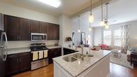 This single-floor residence offers a thoughtfully designed open-concept layout that seamlessly connects the kitchen, dining, and living areas. The kitchen is a standout feature, showcasing rich dark-espresso cabinetry, granite countertops, a subway tile backsplash, and a full suite of stainless steel appliances including a refrigerator, range, microwave, and dishwasher. A large peninsula with an undermount double sink and pendant lighting above provides ample seating and serves as an ideal gathering point. Warm hardwood flooring flows throughout the main living areas, complementing the bright, neutral color palette. The living room is bathed in natural light from large windows and a glass door, creating an inviting and airy atmosphere. Two bathrooms continue the home's cohesive design with granite-topped dark wood vanities and tile flooring; one features a walk-in shower, while the other offers a tub-shower combination. The bedrooms are carpeted for comfort, with generous windows providing natural light. One additional room with carpet and dual windows offers flexible use as a bedroom, office, or flex space. Throughout the residence, clean architectural lines, high ceilings in the kitchen and entry area, and consistent high-quality finishes contribute to a polished and comfortable living environment.
