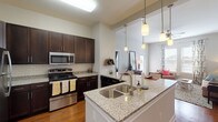 This single-floor residence offers a thoughtfully designed open-concept layout that seamlessly connects the kitchen, dining, and living areas. The kitchen is a standout feature, showcasing rich dark-espresso cabinetry, granite countertops, a subway tile backsplash, and a full suite of stainless steel appliances including a refrigerator, range, microwave, and dishwasher. A large peninsula with an undermount double sink and pendant lighting above provides ample seating and serves as an ideal gathering point. Warm hardwood flooring flows throughout the main living areas, complementing the bright, neutral color palette. The living room is bathed in natural light from large windows and a glass door, creating an inviting and airy atmosphere. Two bathrooms continue the home's cohesive design with granite-topped dark wood vanities and tile flooring; one features a walk-in shower, while the other offers a tub-shower combination. The bedrooms are carpeted for comfort, with generous windows providing natural light. One additional room with carpet and dual windows offers flexible use as a bedroom, office, or flex space. Throughout the residence, clean architectural lines, high ceilings in the kitchen and entry area, and consistent high-quality finishes contribute to a polished and comfortable living environment.