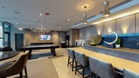 Located at 600 S Wells St in Chicago's vibrant South Loop neighborhood, this sophisticated amenity lounge is designed to offer residents a comprehensive social and entertainment destination all on one level. The space is divided into several distinct yet open-concept zones, each thoughtfully appointed with contemporary furnishings and upscale finishes throughout.

The game room area anchors one end of the space, featuring a full-size billiards table beneath a striking multi-arm chandelier, a wall-mounted flat-screen TV, and textured accent wall panels that add warmth and visual depth. An adjacent bar and kitchen area is outfitted with sleek, floor-to-ceiling cabinetry in a neutral wood tone, a dark stone backsplash, integrated appliances, and a generous counter-height island with leather barstools — all illuminated by elegant pendant lighting.

The media lounge offers generous seating arrangements centered around a large-format video wall, creating an immersive viewing experience. Nearby, a quieter lounge nook provides plush armchairs and curated wall art, including a built-in board game display. Floor-to-ceiling glass walls and sliding doors connect the interior to an outdoor terrace, flooding the space with natural light and offering a seamless indoor-outdoor flow. Recessed lighting, patterned carpet, and refined décor unify the entire space with a polished, modern aesthetic.